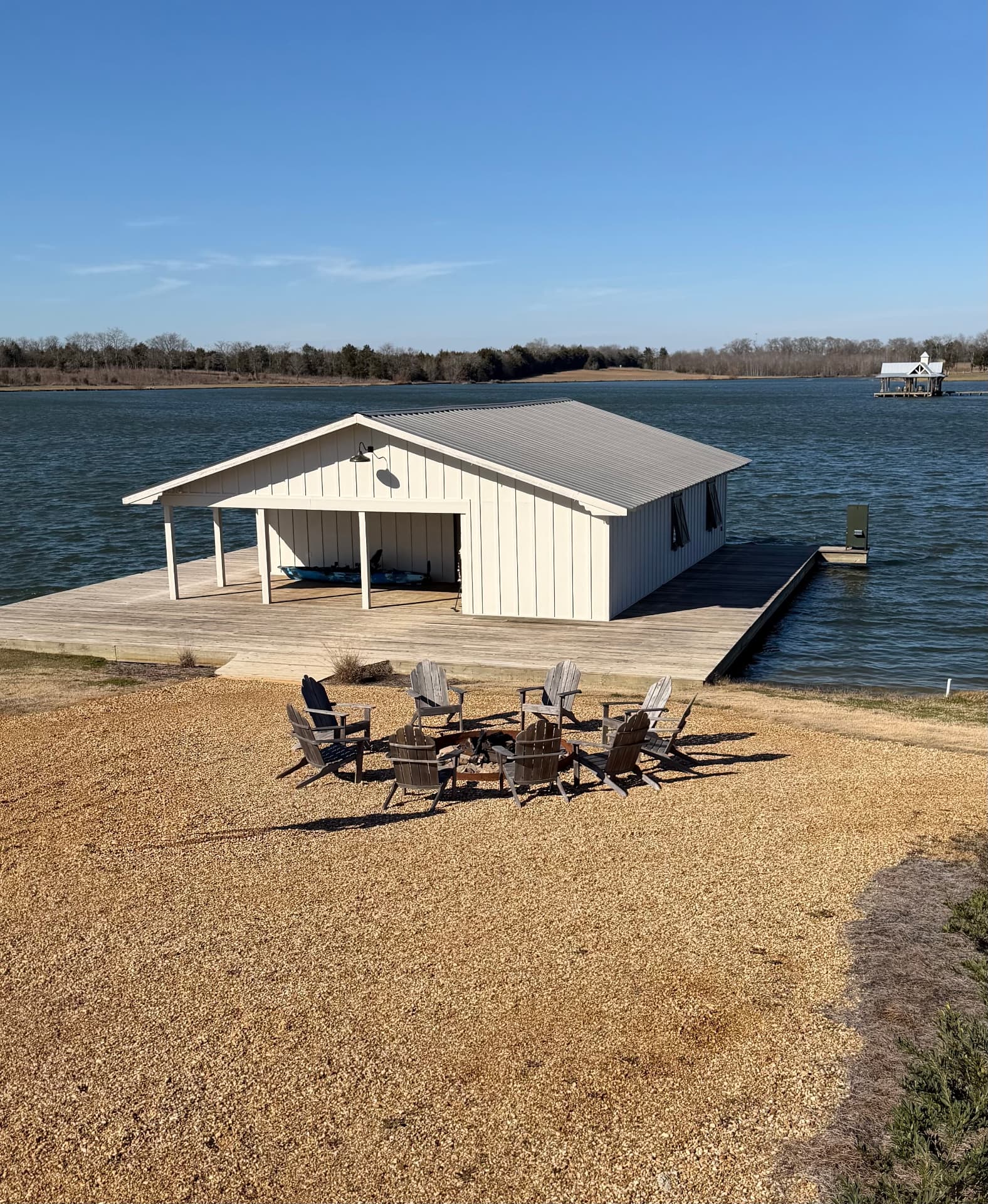White board-and-batten boathouse on a wooden dock at Lake Faunsdale, with a gravel fire-pit patio in front and open water beyond